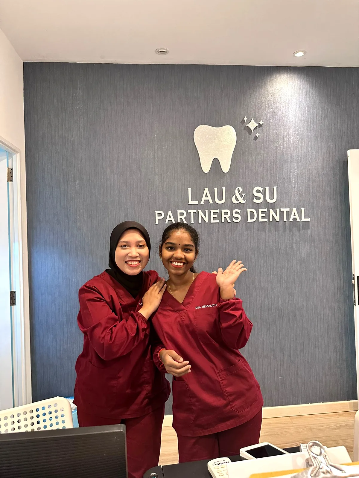 Two dental nurses in maroon scrubs smiling at the reception desk in front of the Lau & Su Partners Dental sign in Bukit Jalil