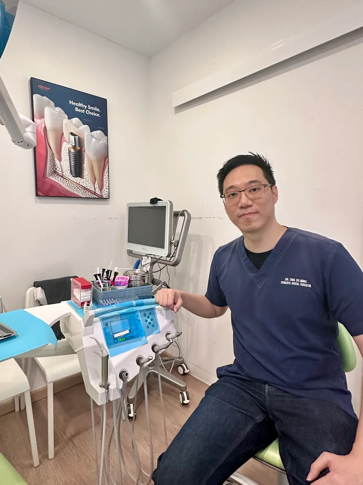 Dr. Ong De Ming, resident dental surgeon, seated beside the dental chair and instrument cart in a treatment room at Lau & Su Partners Dental Bukit Jalil