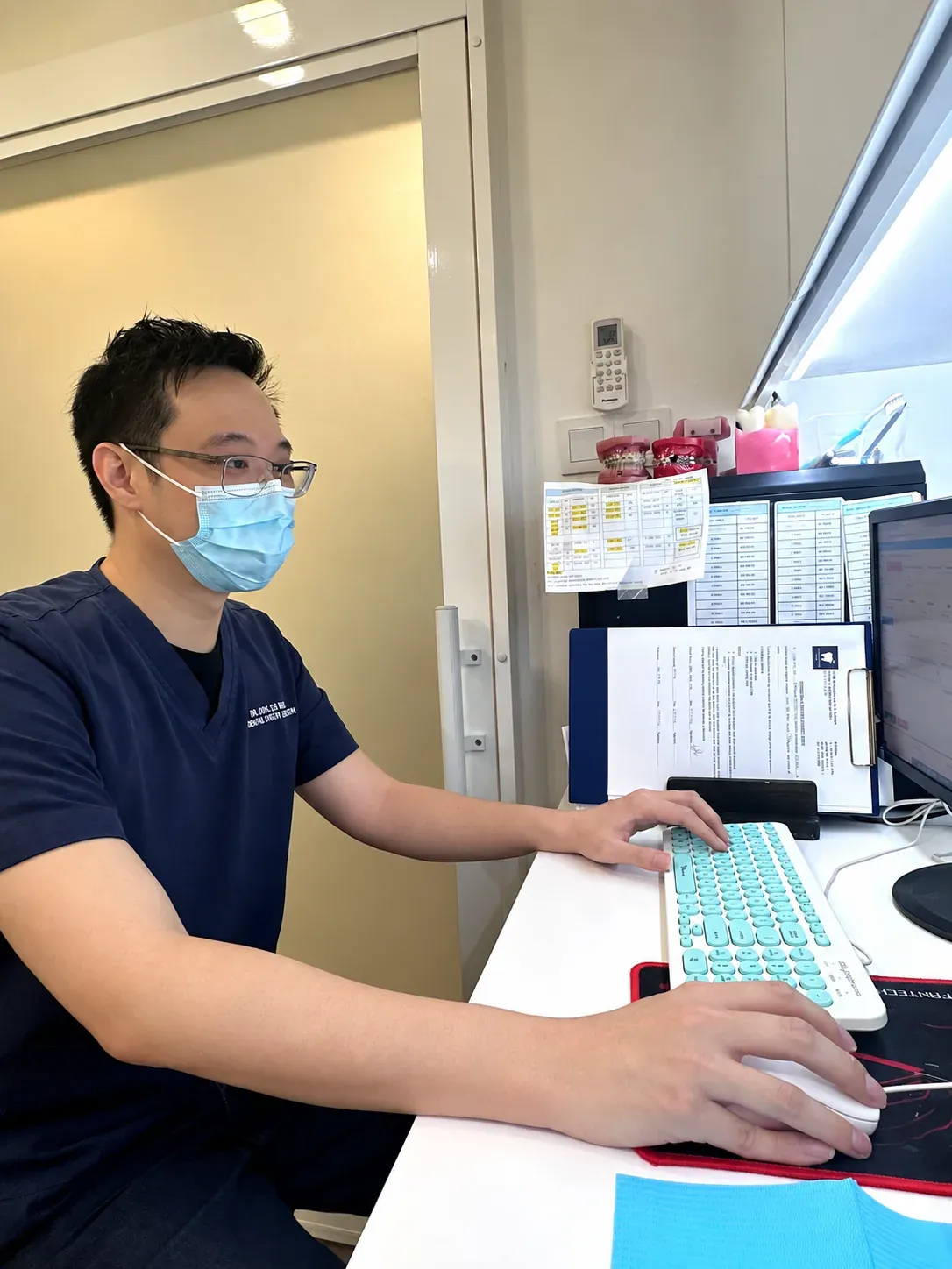 Dr. Ong De Ming in a face mask updating patient records at the computer between appointments at Lau & Su Partners Dental Bukit Jalil