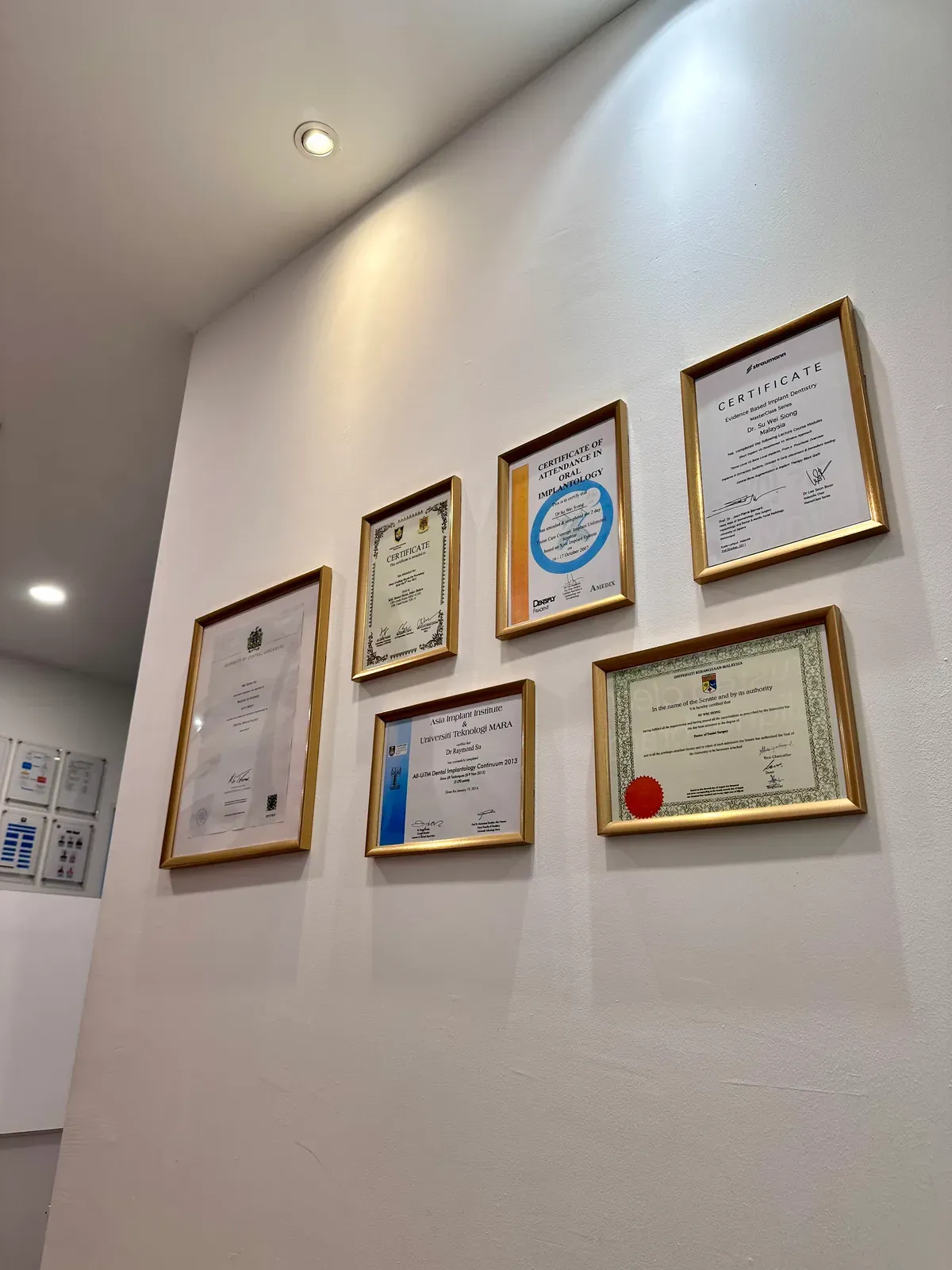 Wall of framed dental degrees, memberships and certificates held by the dentists at Lau & Su Partners Dental Bukit Jalil